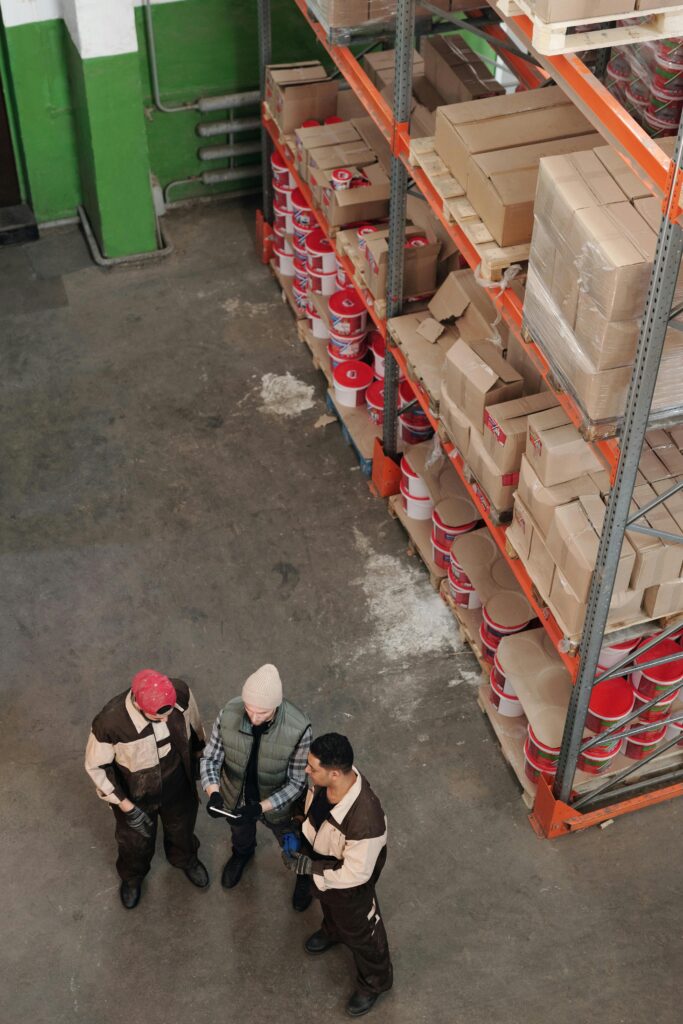 Kariera Group of warehouse workers discussing logistics and inventory in a large storage area with shelves and packages.